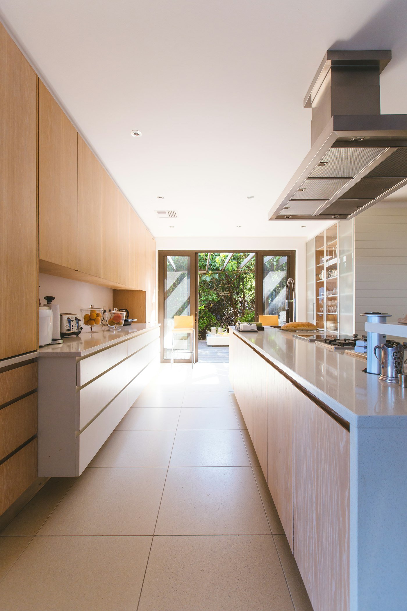 A long kitchen with pale wood cabinets, a stone worktop, and glass doors to a garden